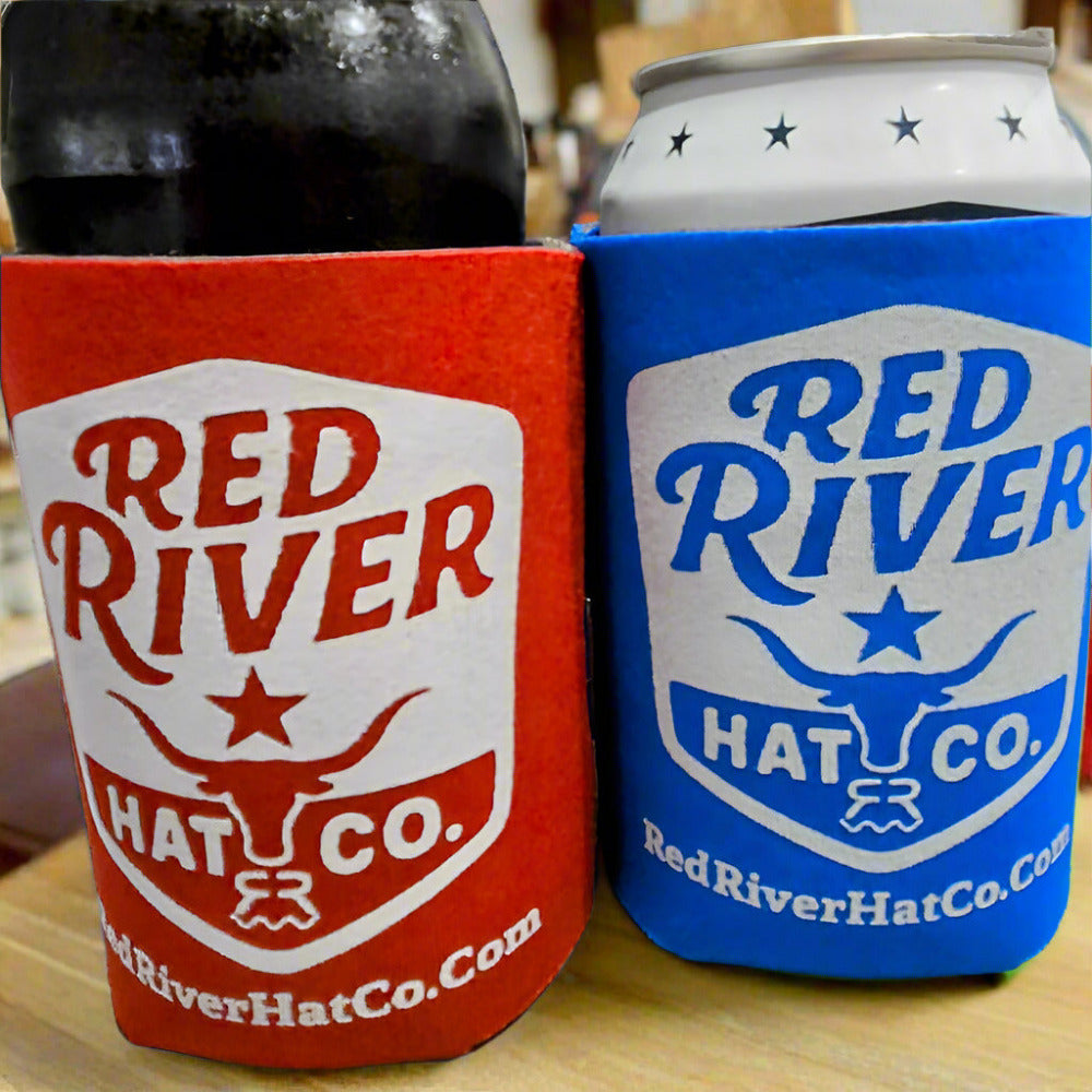 Two Red River Hat Co. branded can coolers, one red and one blue, on a wooden surface.