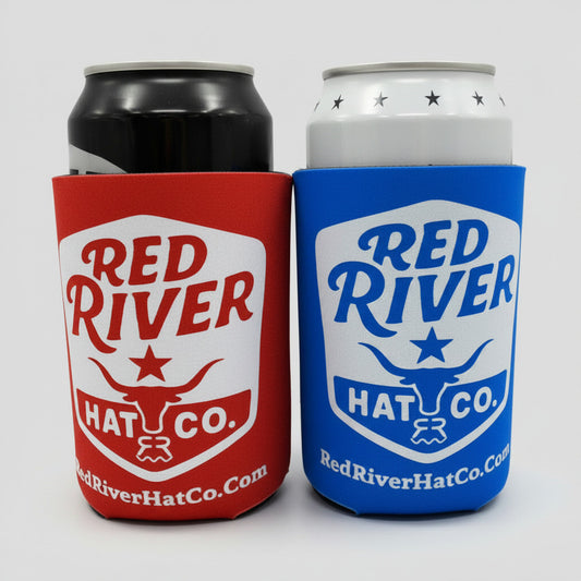 Two Red River Hat Co. branded can coolers, one red and one blue, on a wooden surface.