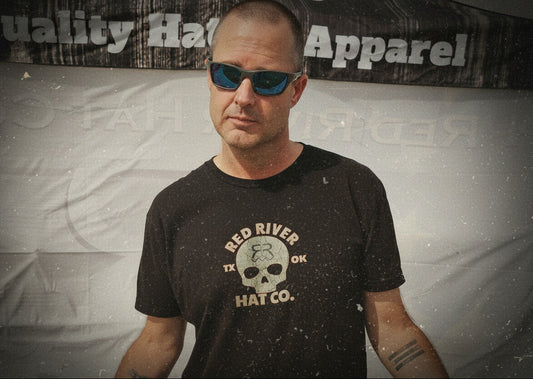 Man wearing sunglasses and a black t-shirt with a logo, standing in front of a banner with text.
