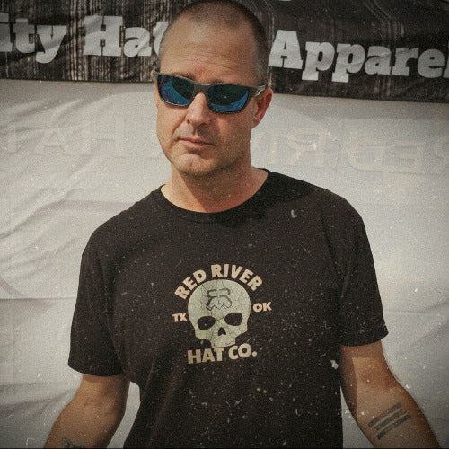 Man wearing sunglasses and a black t-shirt with a logo, standing in front of a banner with text.